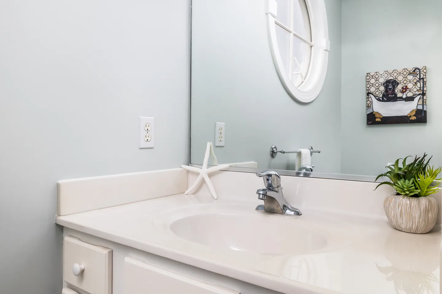 Bathroom vanity with a white countertop, oval sink, chrome faucet, decorative starfish, and reflection of a round window in the mirror.