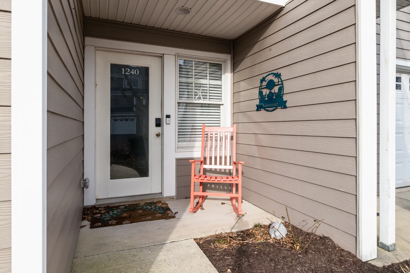 Front entrance of a home featuring a white door with a window, a pink rocking chair, horizontal siding, and a decorative wall sign.