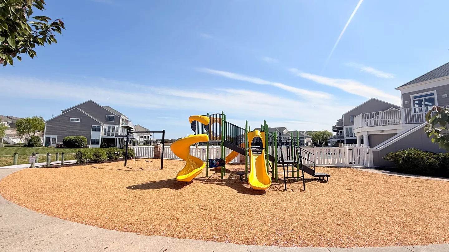 Community playground with yellow slides, green climbing structures, swings, and a wood chip ground cover surrounded by residential townhomes under a blue sky.
