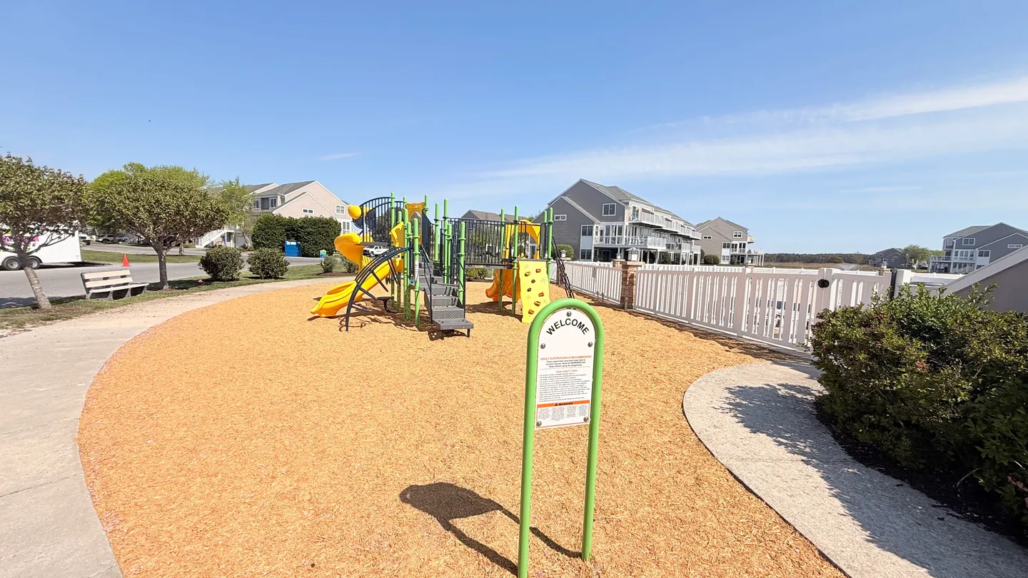 Outdoor community playground with yellow and green play equipment, slides, and wood chip ground cover under a blue sky.