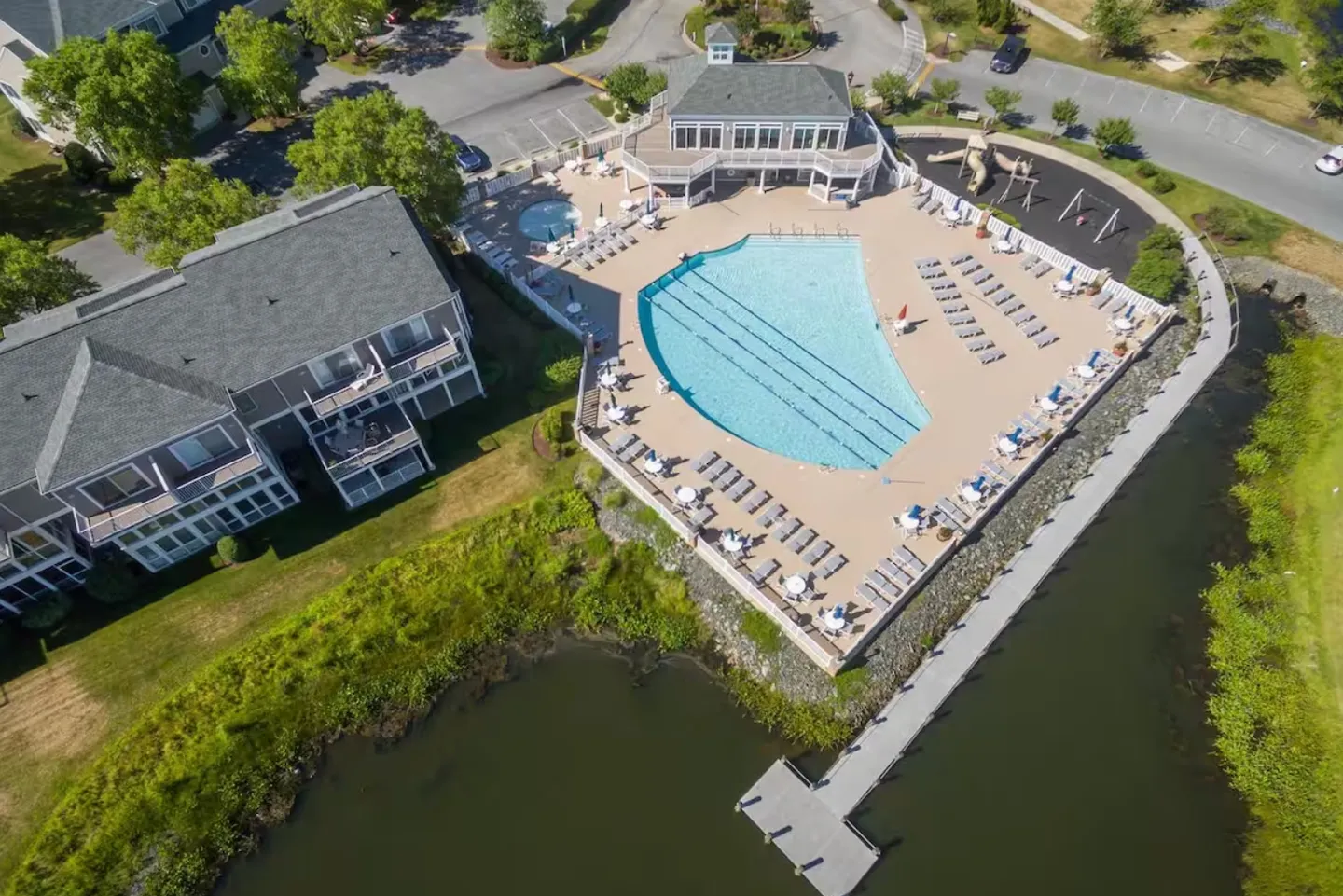 Aerial view of a community swimming pool complex beside a lake, with a clubhouse, sun deck with lounge chairs, and a dock.