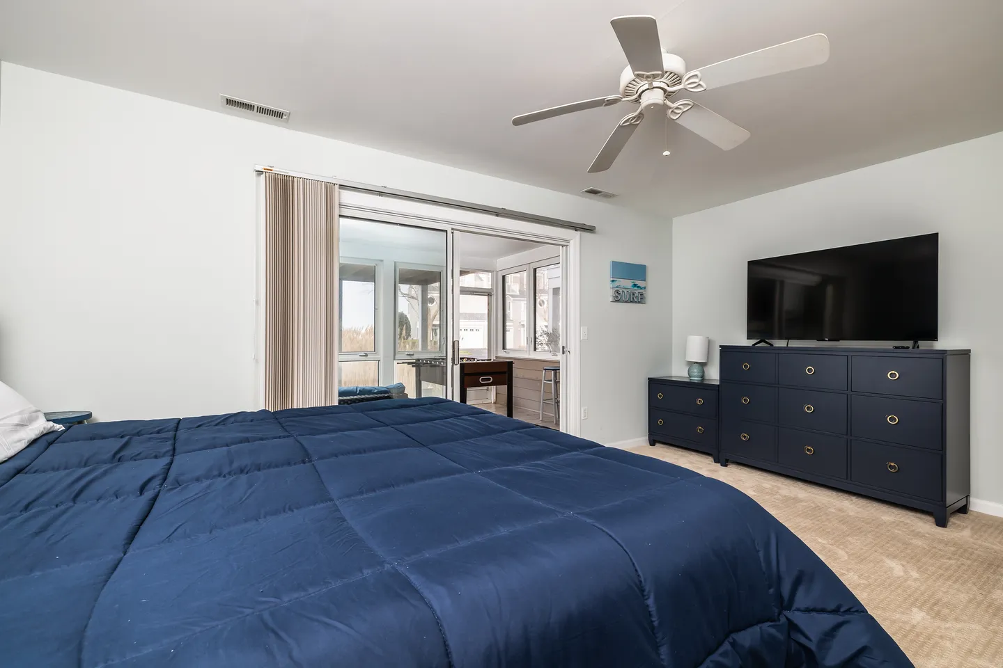 Bedroom with blue bedding, ceiling fan, dark blue dresser with television, and sliding glass door leading to a sunroom.