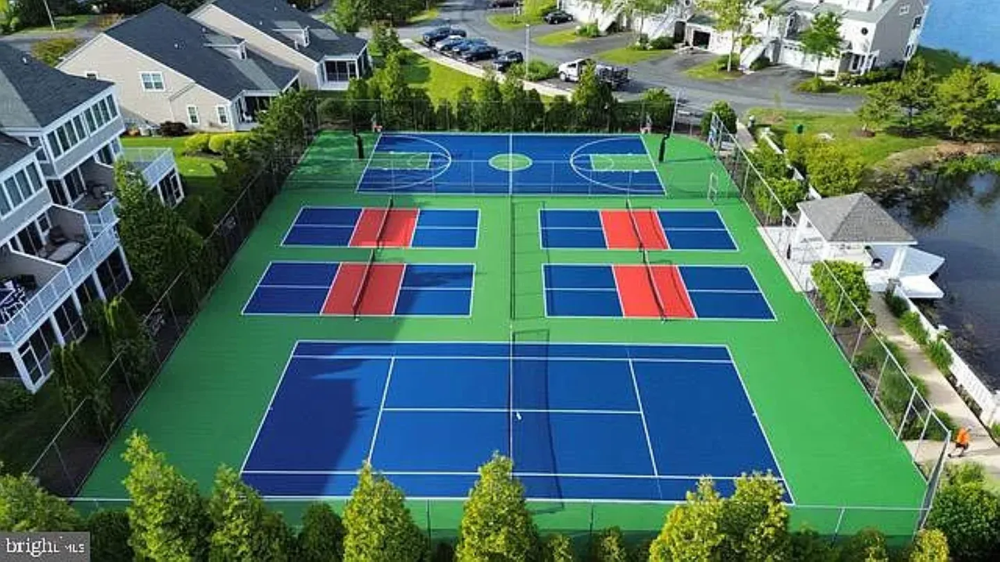 Aerial view of an outdoor sports complex featuring a tennis court, four pickleball courts, and a basketball court next to a pond and residential buildings.
