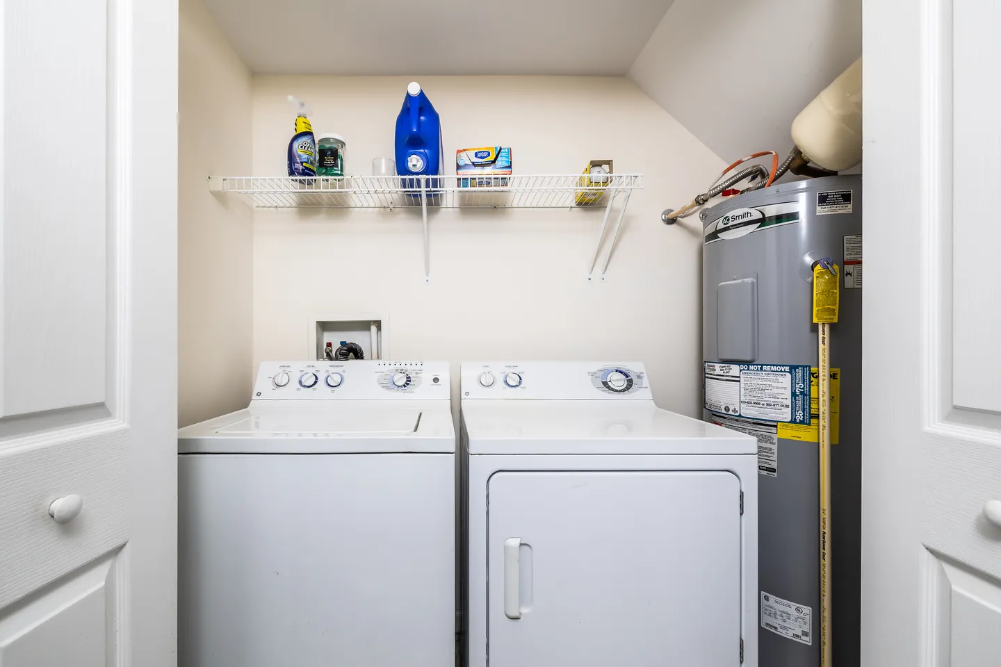 A laundry closet featuring a white washing machine, white dryer, an overhead wire shelf with detergent, and a tall water heater.