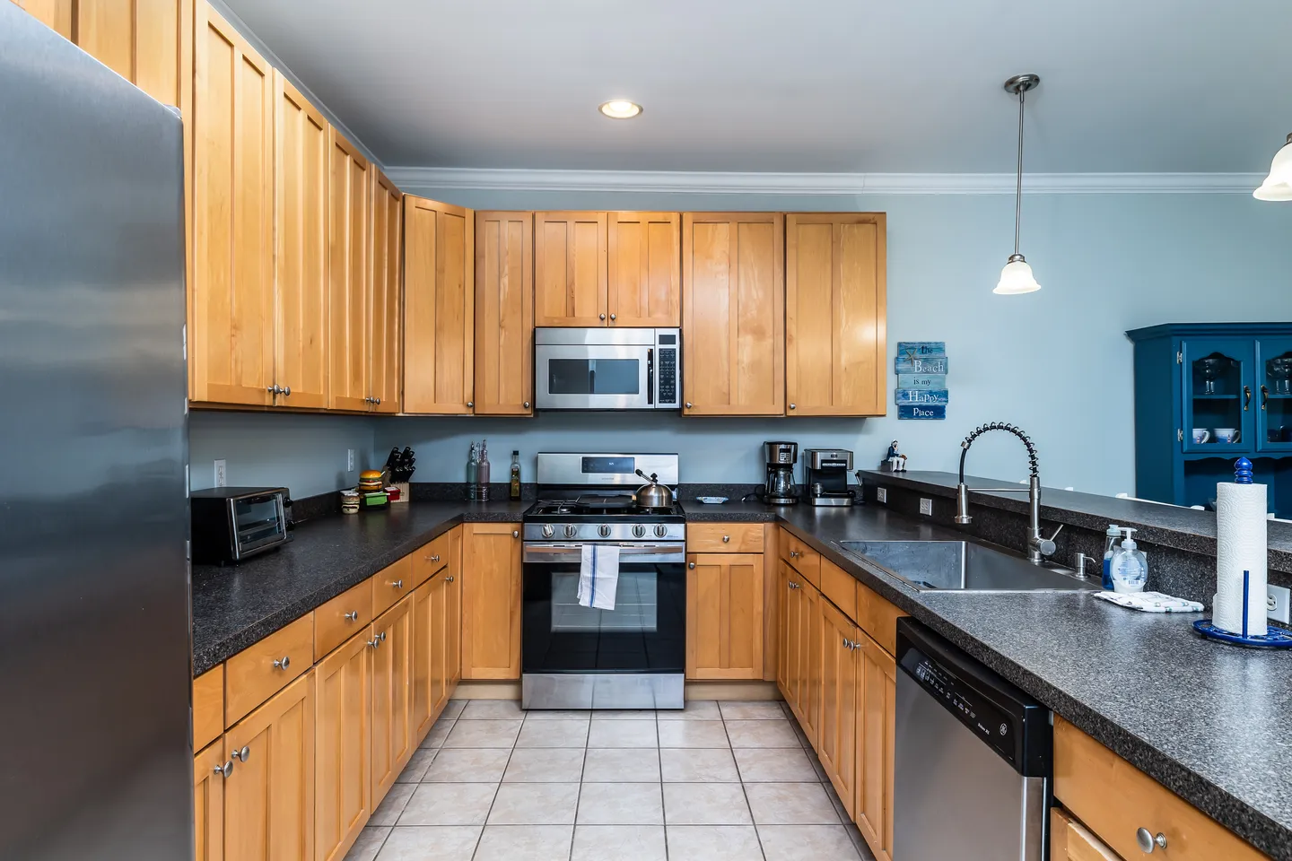 A kitchen with light wood cabinets, dark countertops, tile flooring, and stainless steel appliances.