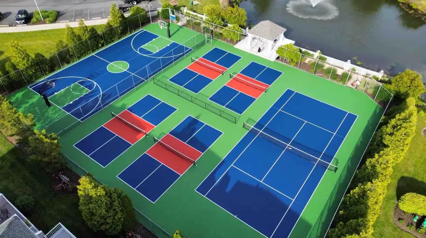 Aerial view of an outdoor sports complex featuring a tennis court, pickleball courts, and a basketball court next to a pond with a fountain.