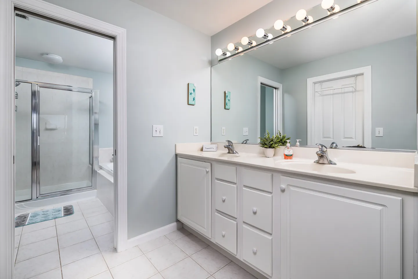 Bathroom with a double-sink white vanity, wide mirror with vanity lights, and a doorway leading to a tiled shower area.