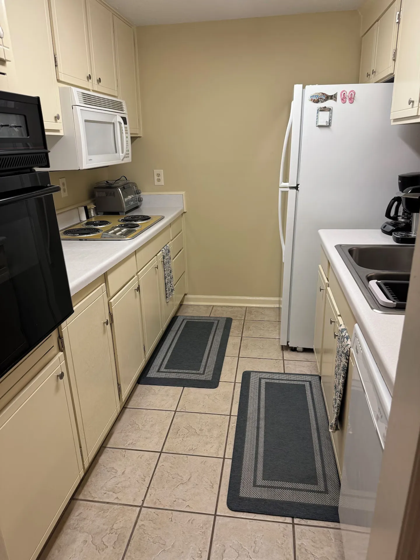 Galley kitchen with cream-colored cabinets, white countertops, built-in wall oven, cooktop, white refrigerator, and tiled floor.