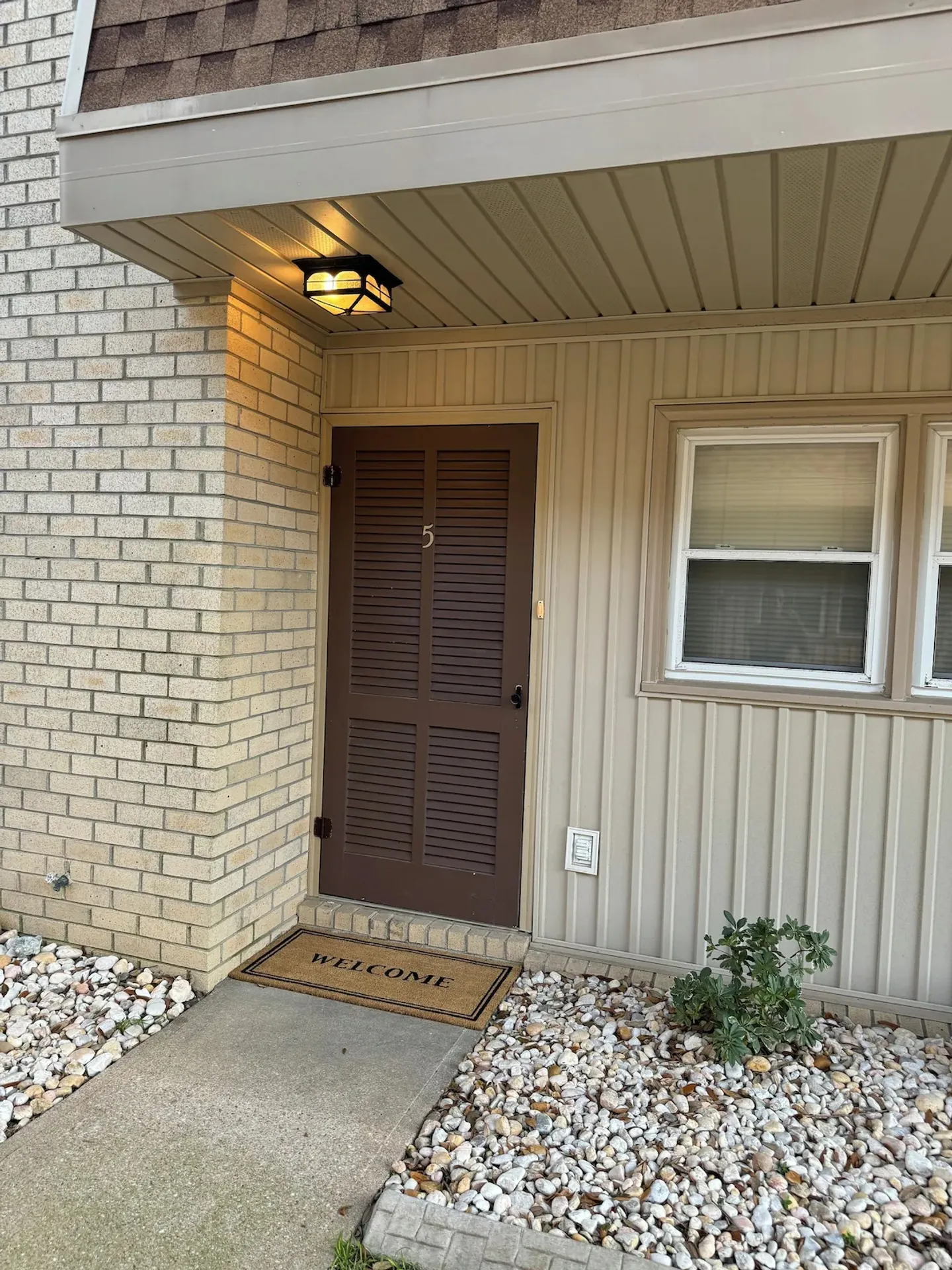 Exterior entrance featuring a brown louvered door numbered 5, a welcome mat, overhead light, and gravel landscaping.