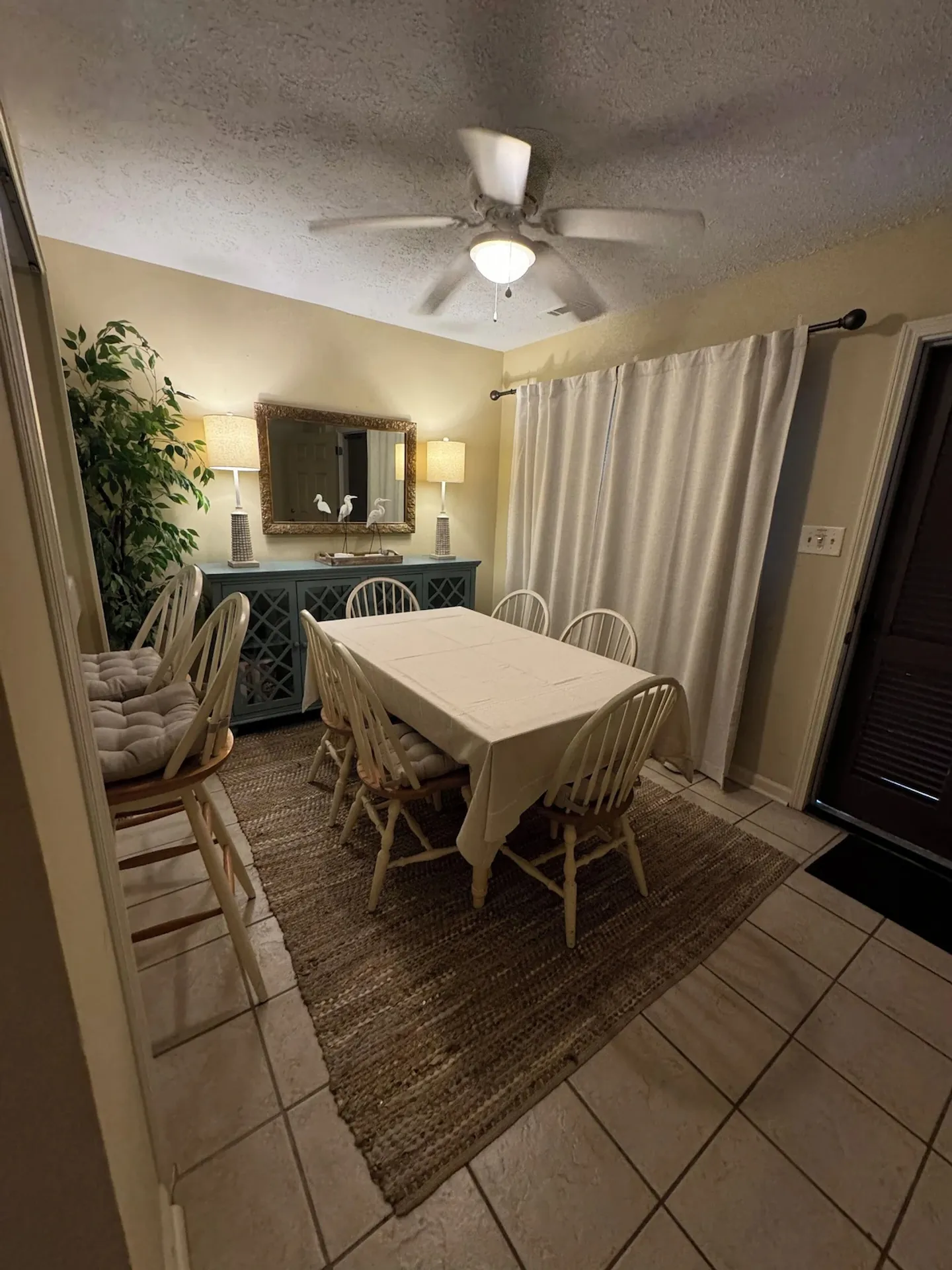 Dining area with a covered table, chairs on a woven rug, a sideboard with lamps, and a ceiling fan overhead.