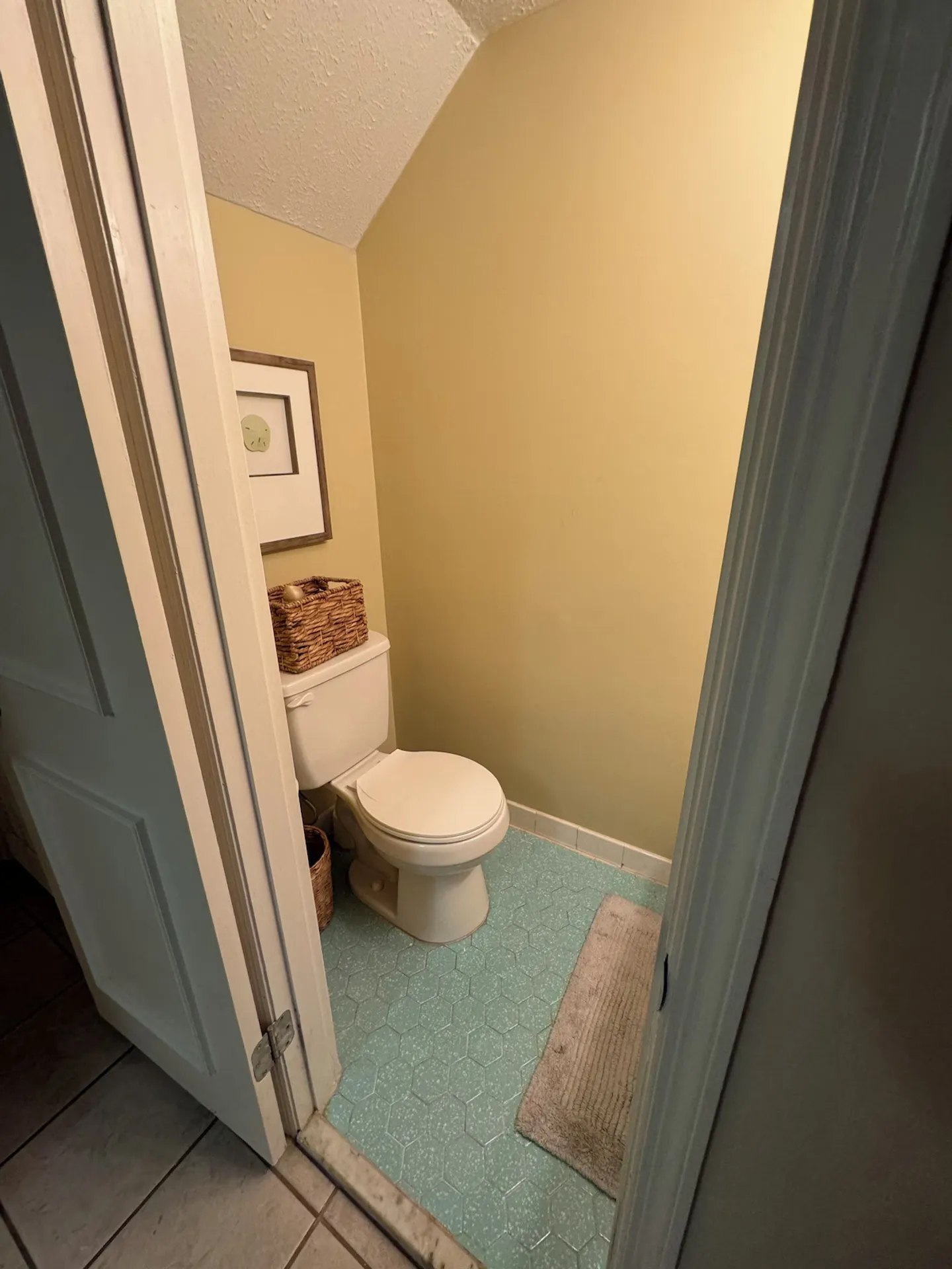 A small water closet with yellow walls, a white toilet, and blue patterned tile flooring viewed through an open doorway.