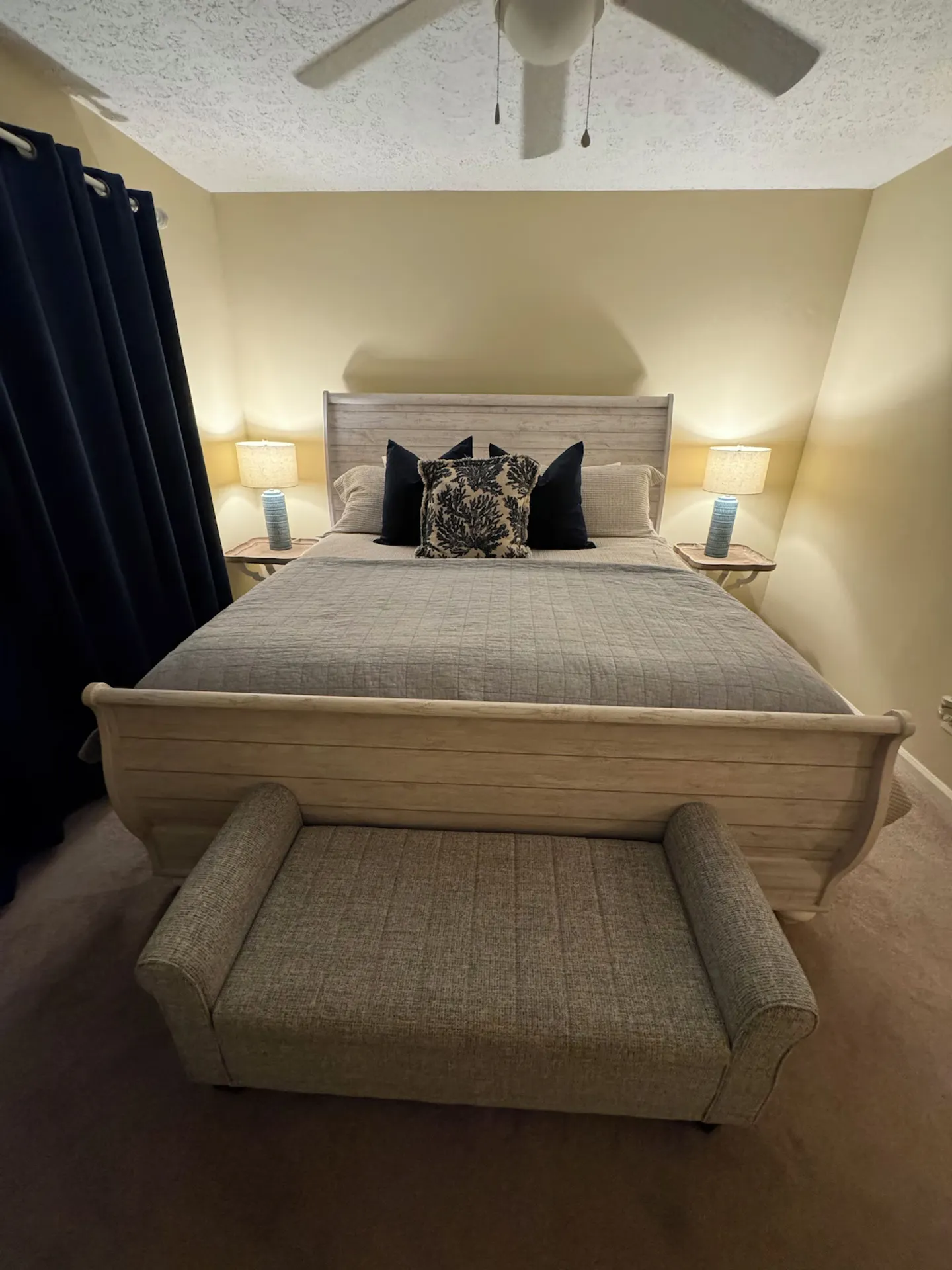 Bedroom featuring a light wood sleigh bed with grey bedding, accent pillows, an upholstered bench at the foot, and two bedside tables with lamps.