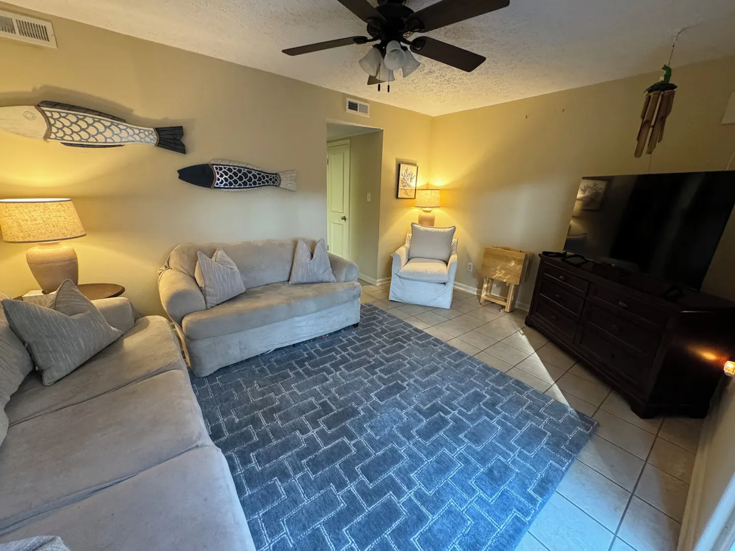 Living room with light-colored sofas, an armchair, tiled flooring, a patterned blue area rug, and wall-mounted fish decor.