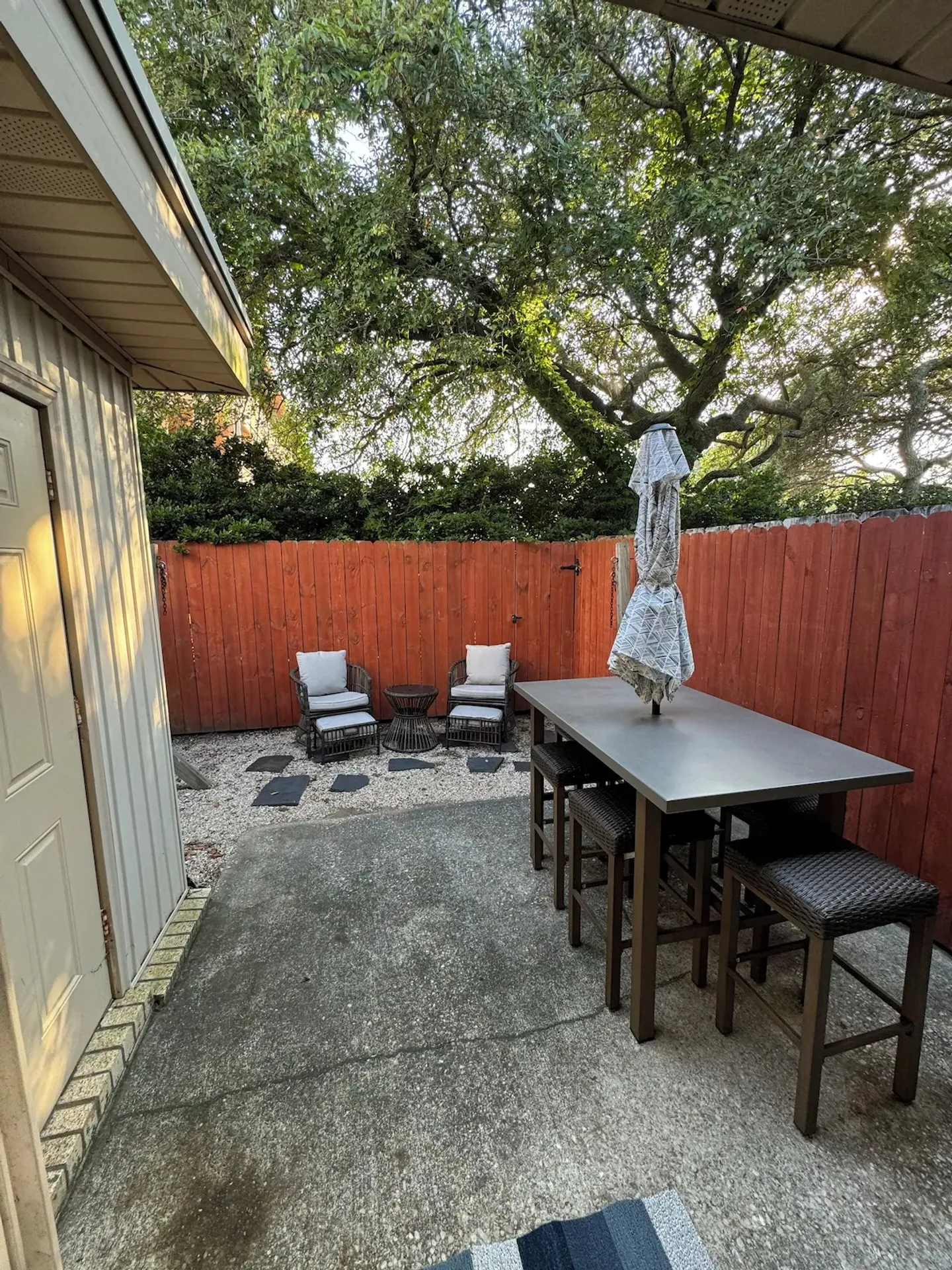 Fenced patio area with outdoor dining table, high-top stools, and a small gravel sitting area with two armchairs.