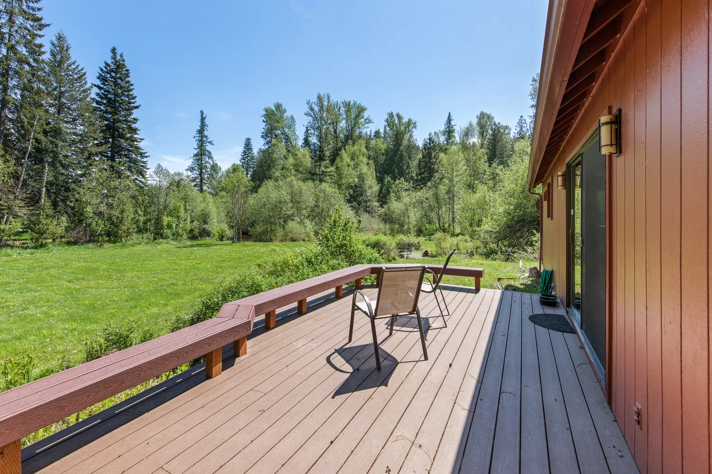 Outdoor wooden deck with bench seating and two chairs overlooking a grassy lawn and dense forest under a clear sky.