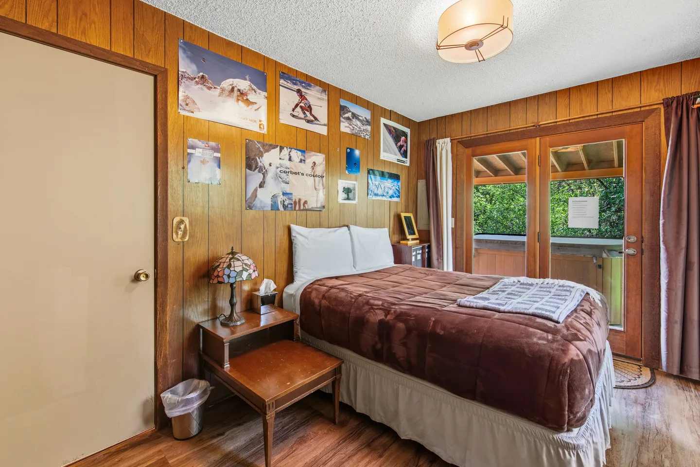Bedroom with wood-paneled walls, a made bed with brown bedding, and sliding glass doors leading outside.