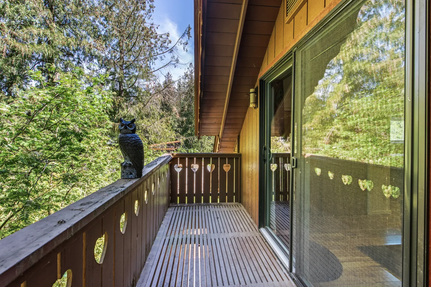 Elevated wooden balcony with heart cutouts on the railing, an owl statue, and sliding glass doors under an overhanging eave overlooking trees.