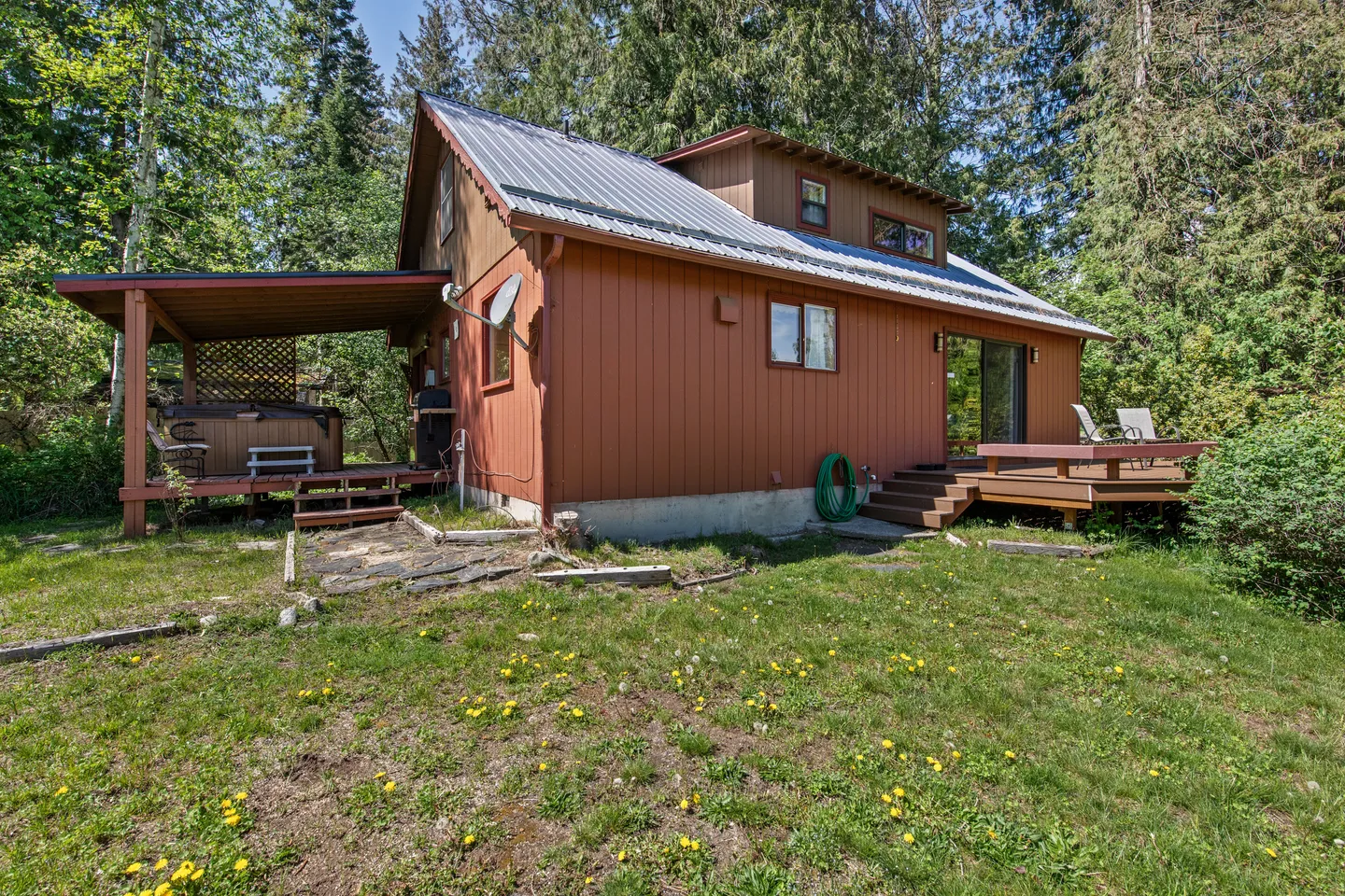 Rear exterior of a brown-sided cabin with a metal roof, open deck, covered porch with hot tub, and grassy yard surrounded by trees.