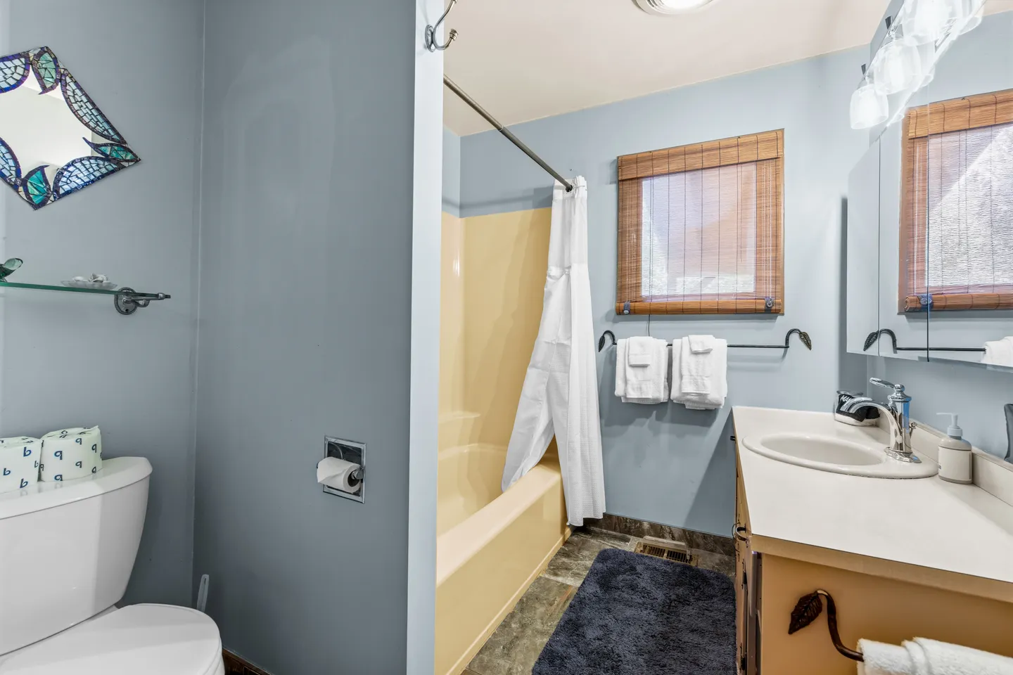Bathroom with light blue walls, vanity sink with mirror, toilet, and yellow bathtub-shower combination with a white curtain.