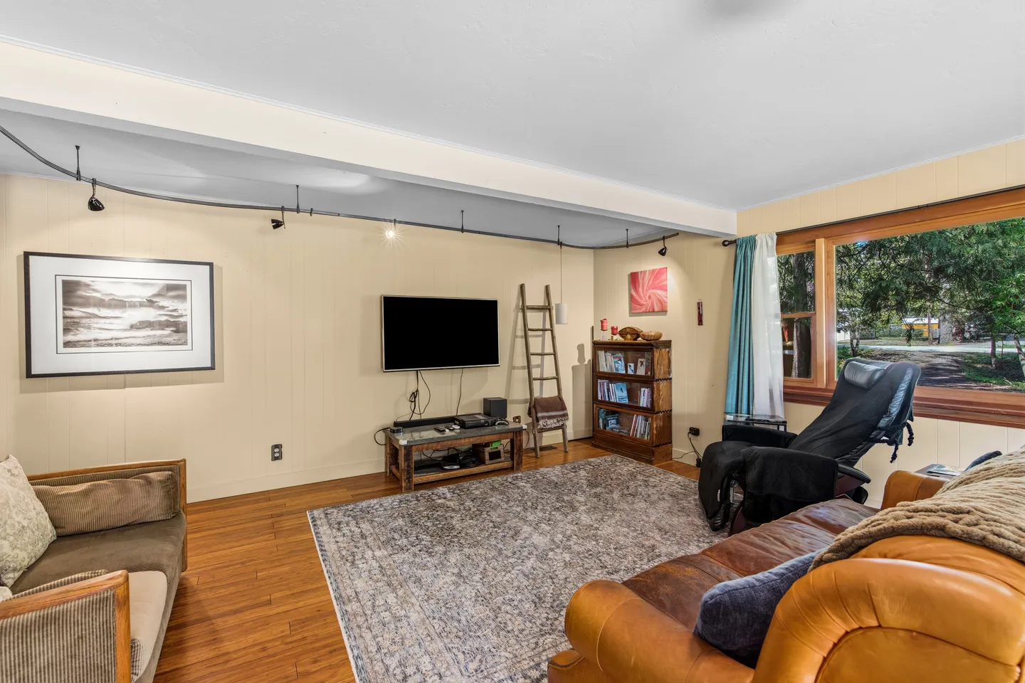 Living room with hardwood floors, an area rug, a wall-mounted TV, leather seating, and track lighting along a ceiling beam.