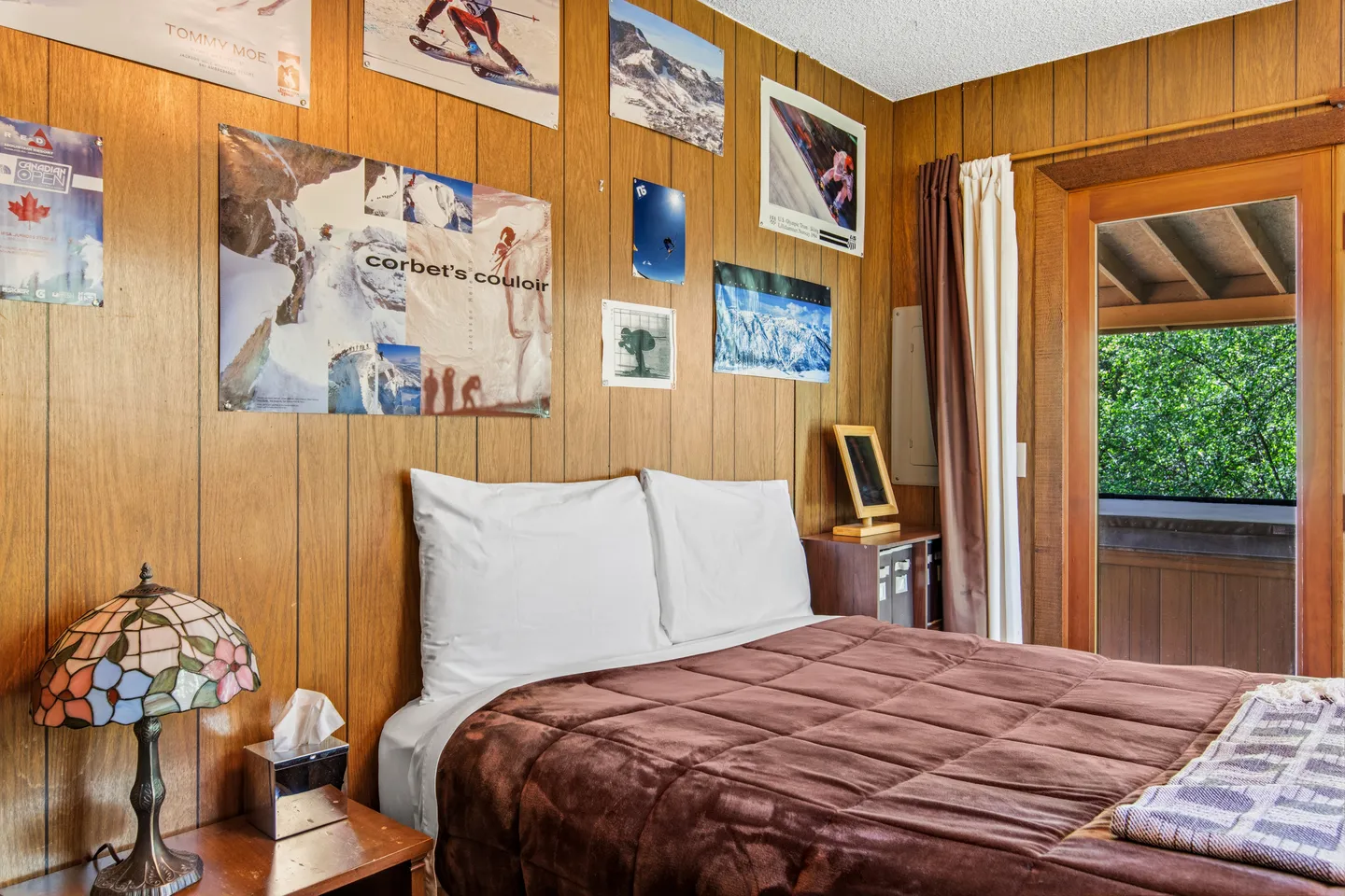 Bedroom with wood-paneled walls, ski posters, a bed with a brown blanket, and a doorway leading outdoors.