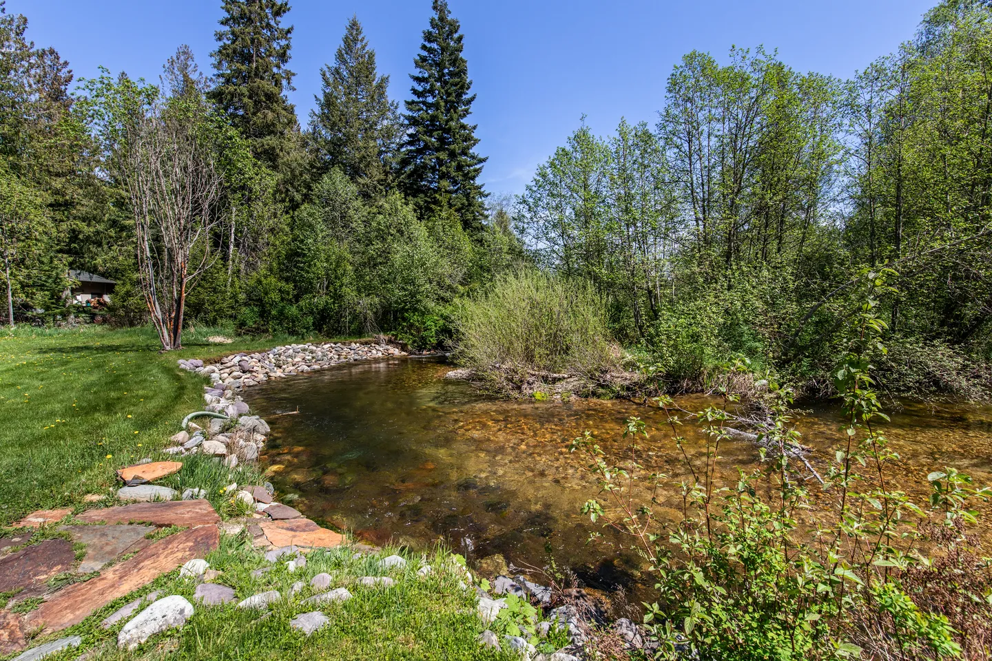 A clear, shallow creek lined with smooth rocks and grass bordering a lush green lawn surrounded by trees under a blue sky.