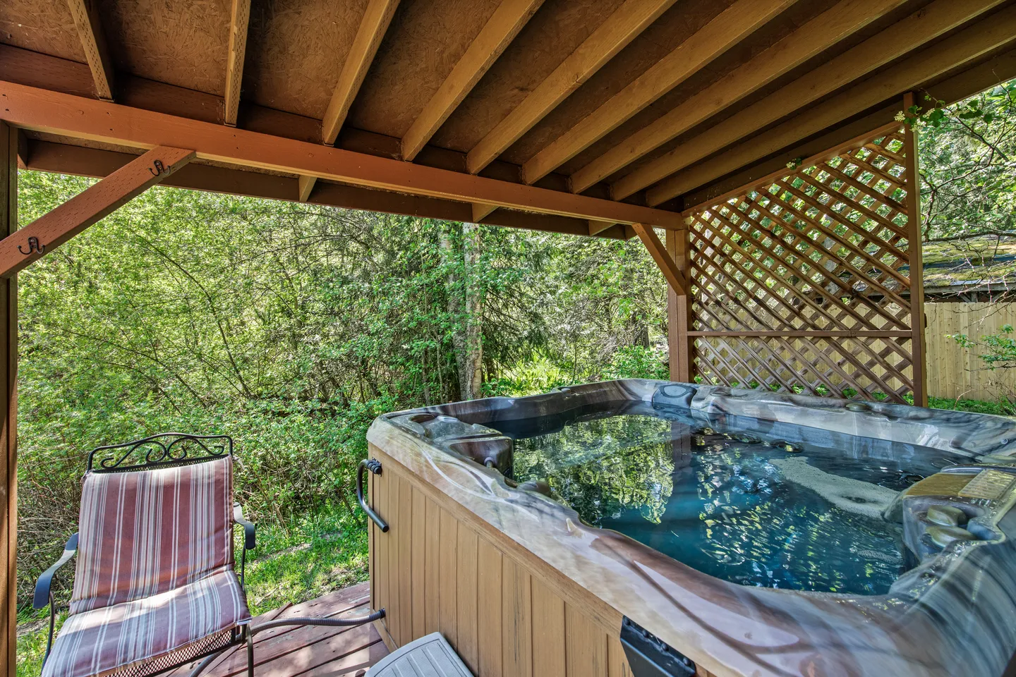 A covered outdoor deck featuring a steaming hot tub with wood siding, a cushioned chair, and a view of surrounding greenery.