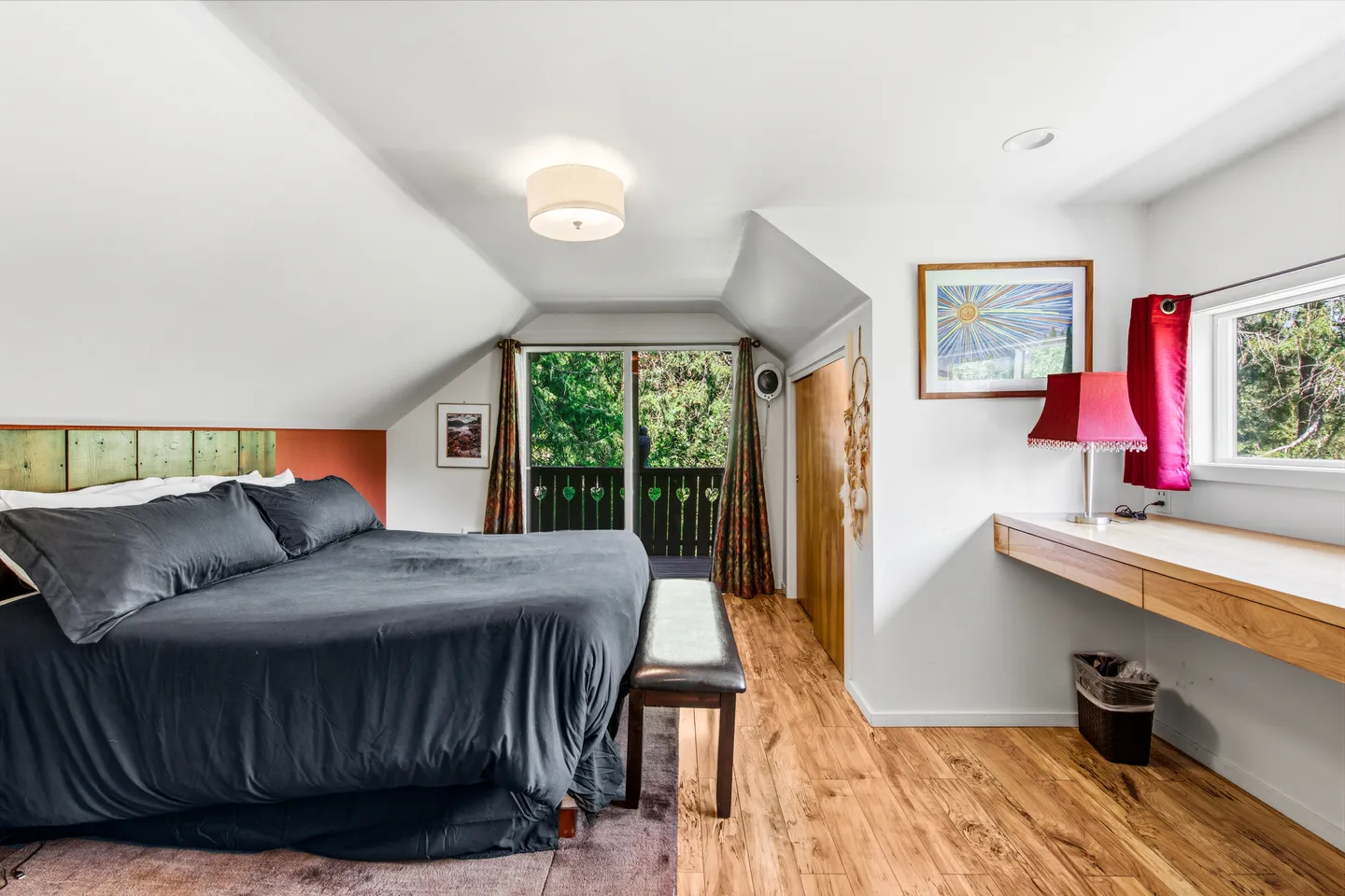Bedroom with angled ceilings, a bed made with dark gray bedding, wood flooring, a built-in desk counter, and glass doors leading to a private balcony.