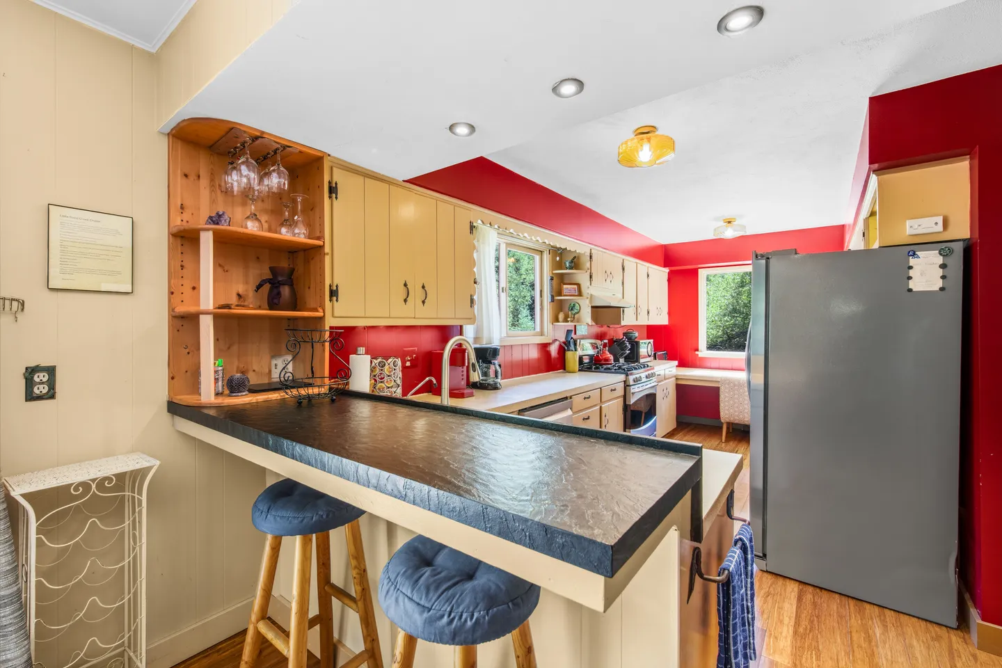 A kitchen with red upper walls, light-colored cabinets, a dark breakfast bar with blue-cushioned wooden stools, and a gray refrigerator.