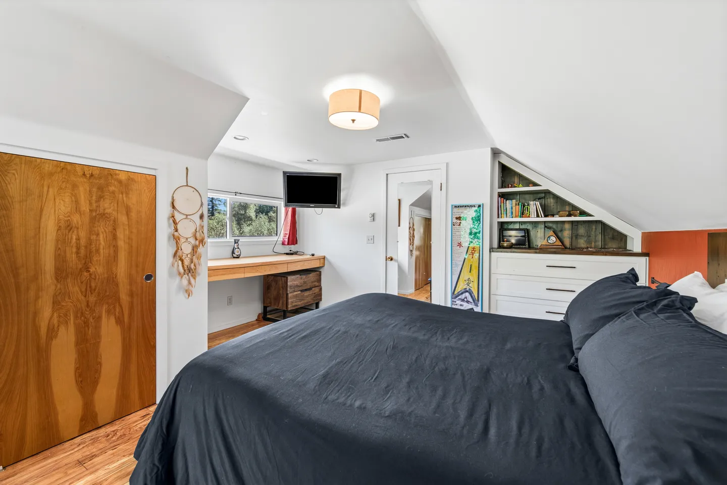 Upstairs bedroom with a sloped ceiling, large bed with dark bedding, built-in dresser, shelves, and a desk area.