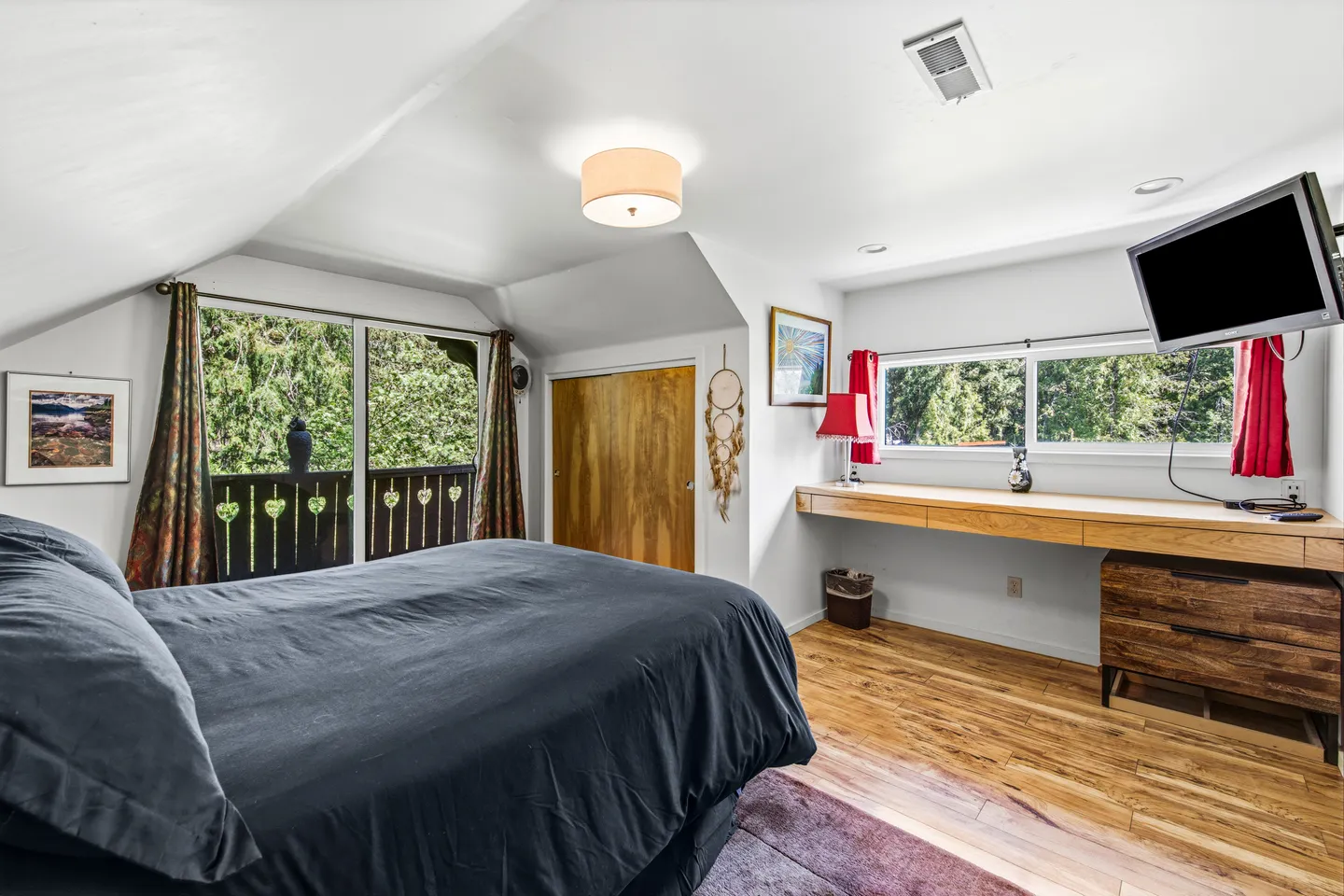 Bedroom with vaulted white ceiling, a large bed with dark bedding, hardwood floors, a long built-in desk, and sliding glass doors leading to a balcony.