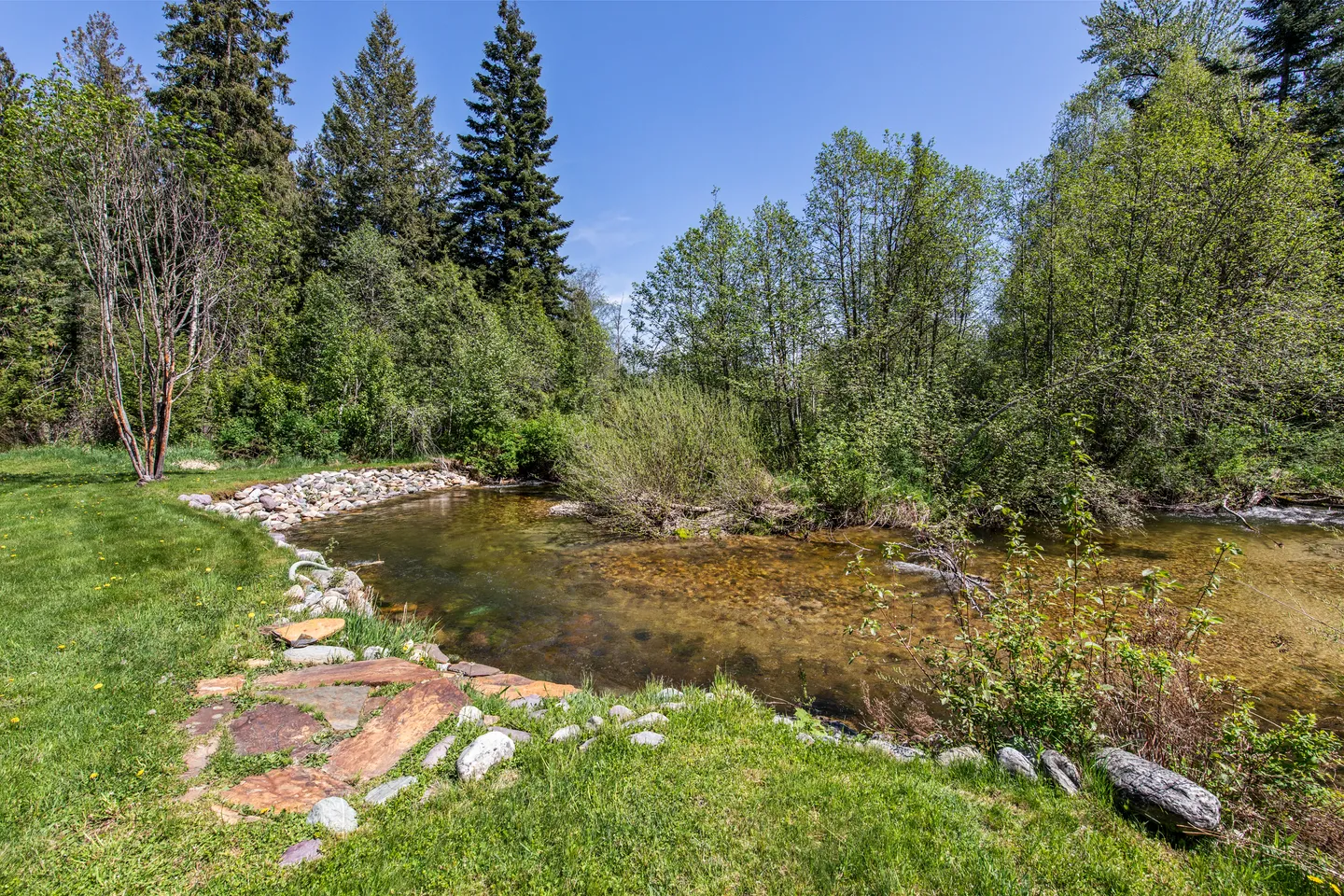 A clear, shallow creek lined with stones and trees bordering a green grassy lawn under a blue sky.