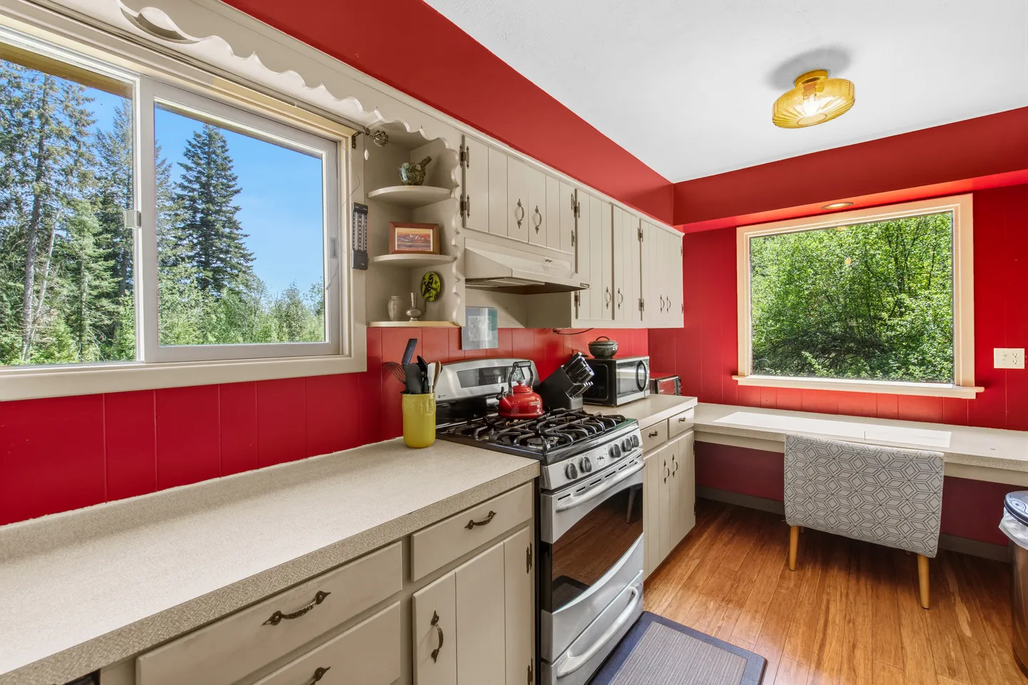 Kitchen with vibrant red walls, light-colored cabinets, a stainless steel stove, and windows overlooking evergreen trees.