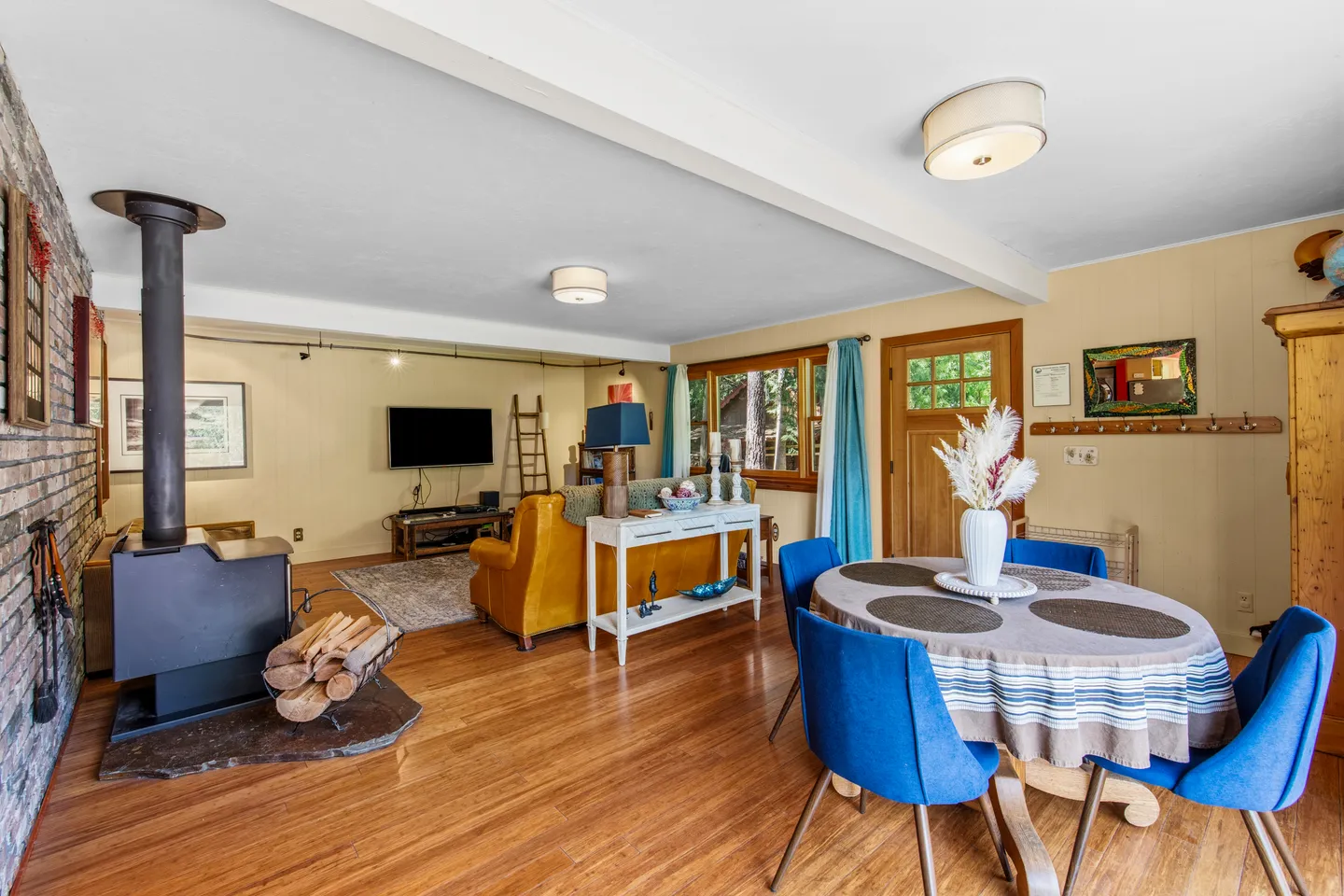 Open-concept living and dining area featuring wood flooring, a dining table with blue chairs, a wood-burning stove, and a sitting area with a sofa and television.