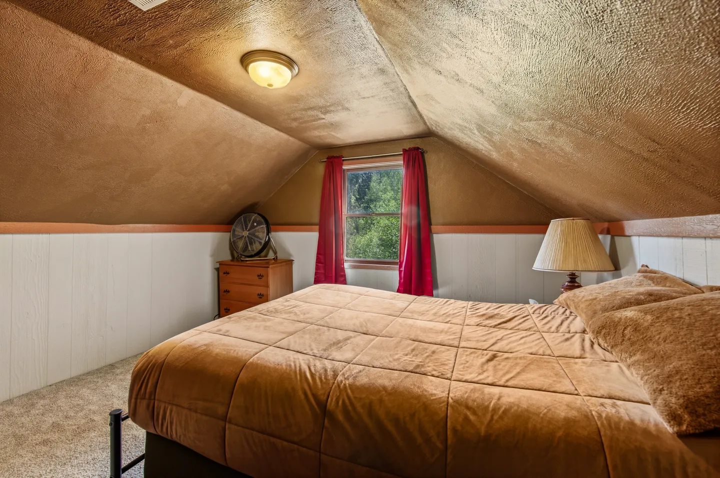 Attic bedroom with sloped ceilings, a made bed with brown bedding, and a small window with red curtains.