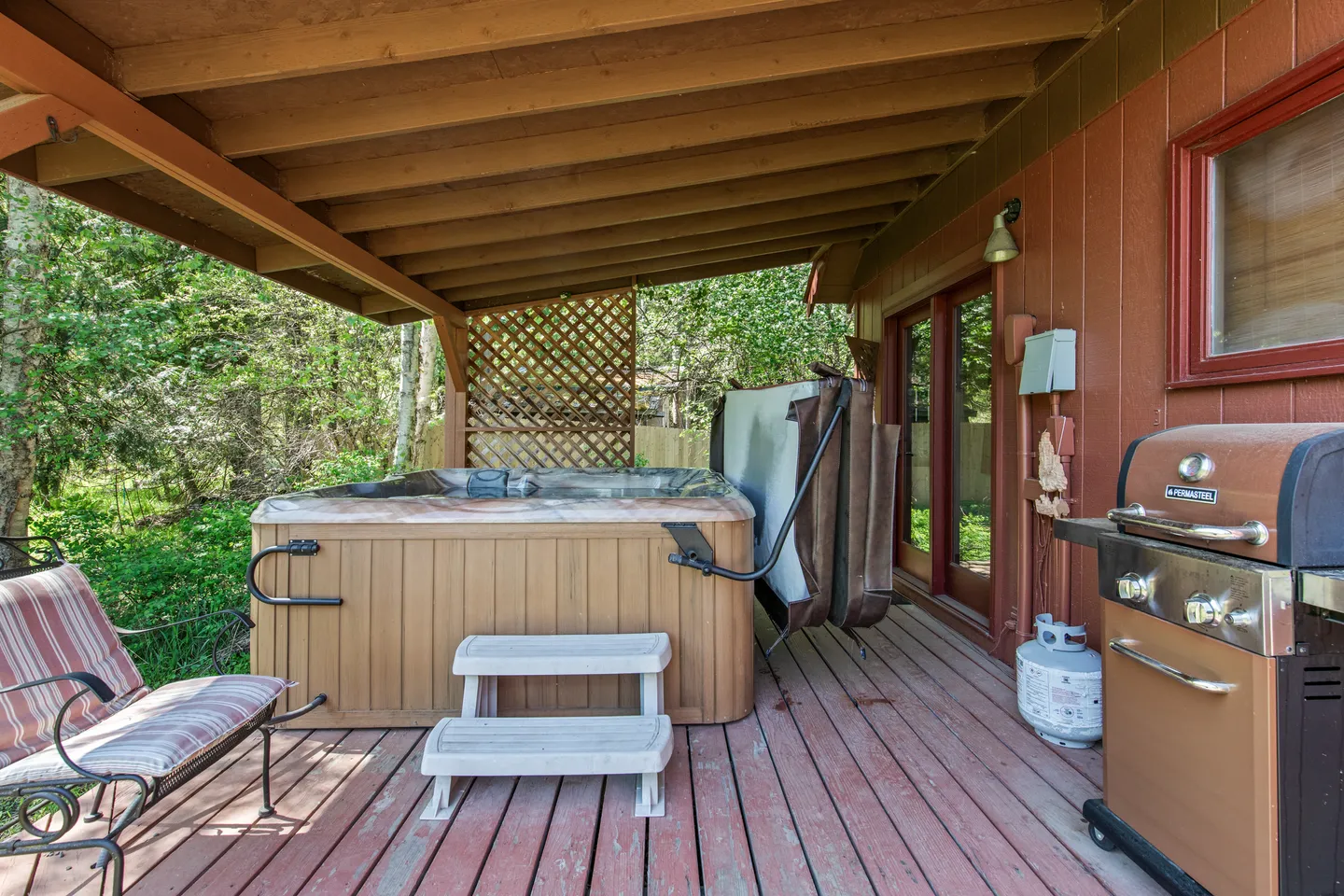 A covered wooden deck with a hot tub, steps, a grill, and an outdoor chair.