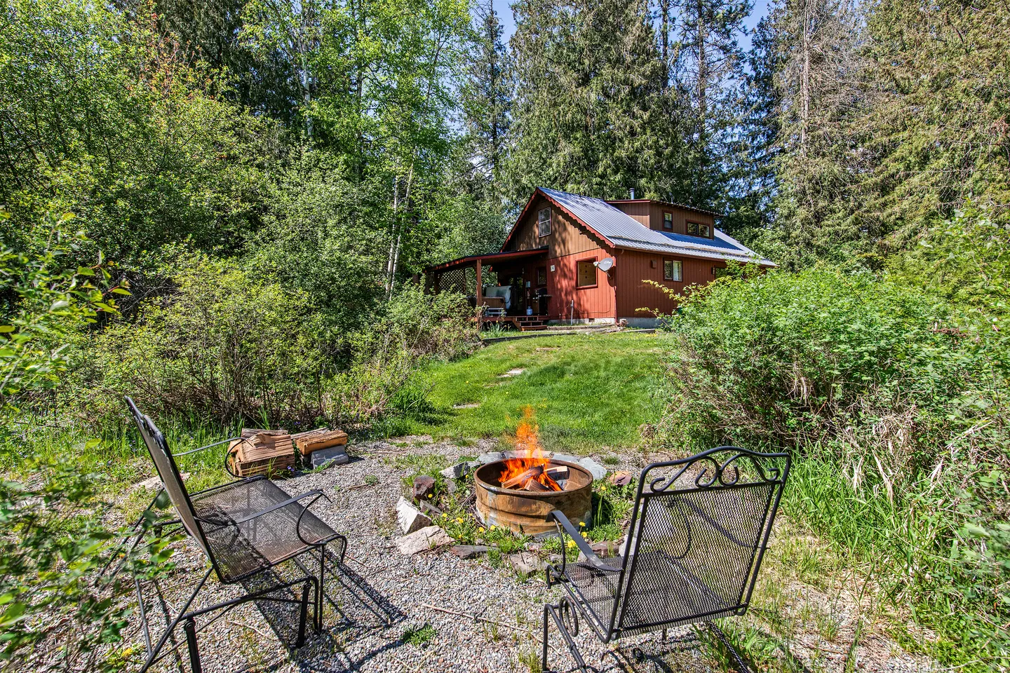 Outdoor fire pit with metal chairs in a gravel area overlooking a lawn and wooden cabin surrounded by trees.