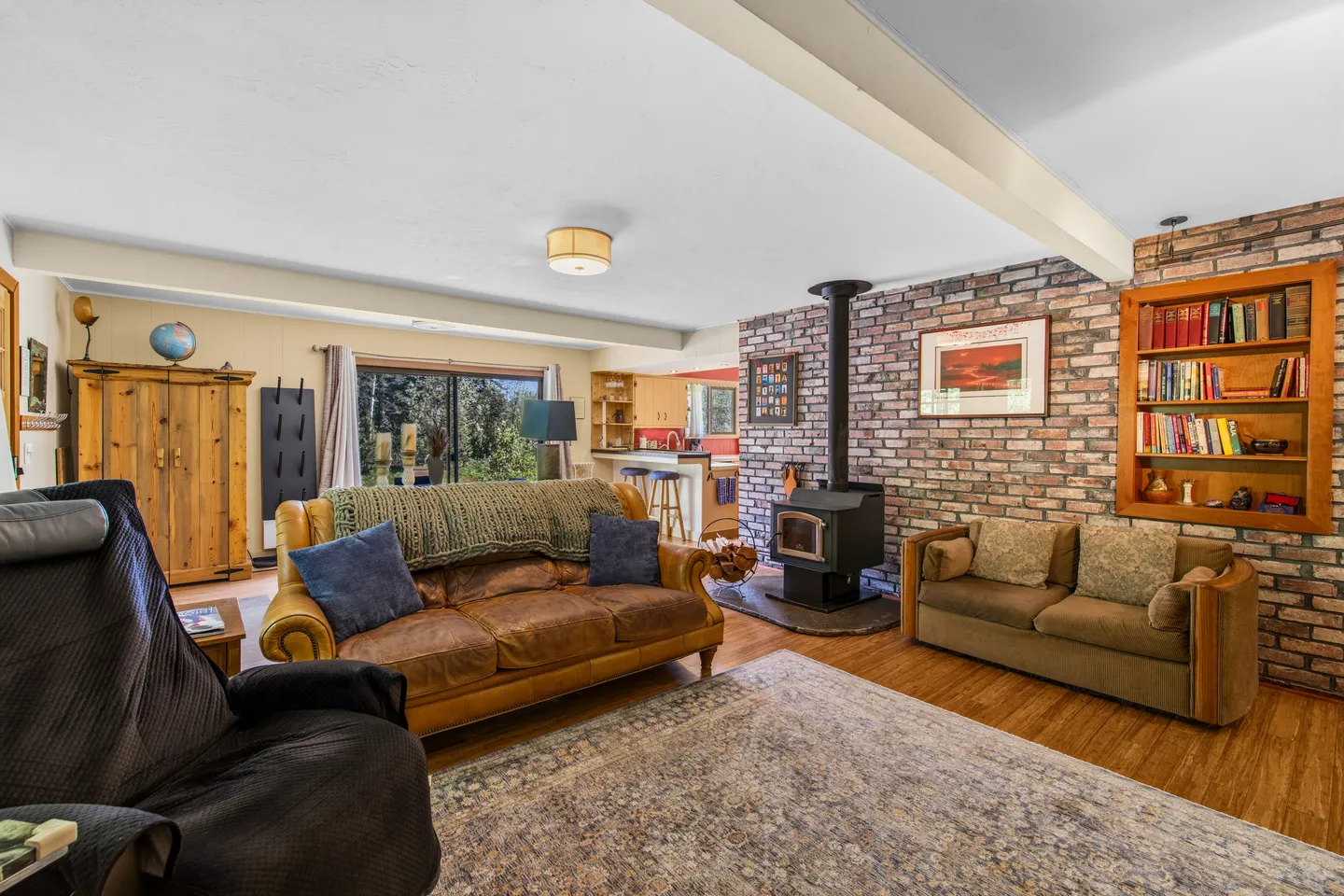 Living room featuring a brick accent wall with a wood stove and built-in bookcase, sofas, and an open view to the kitchen.