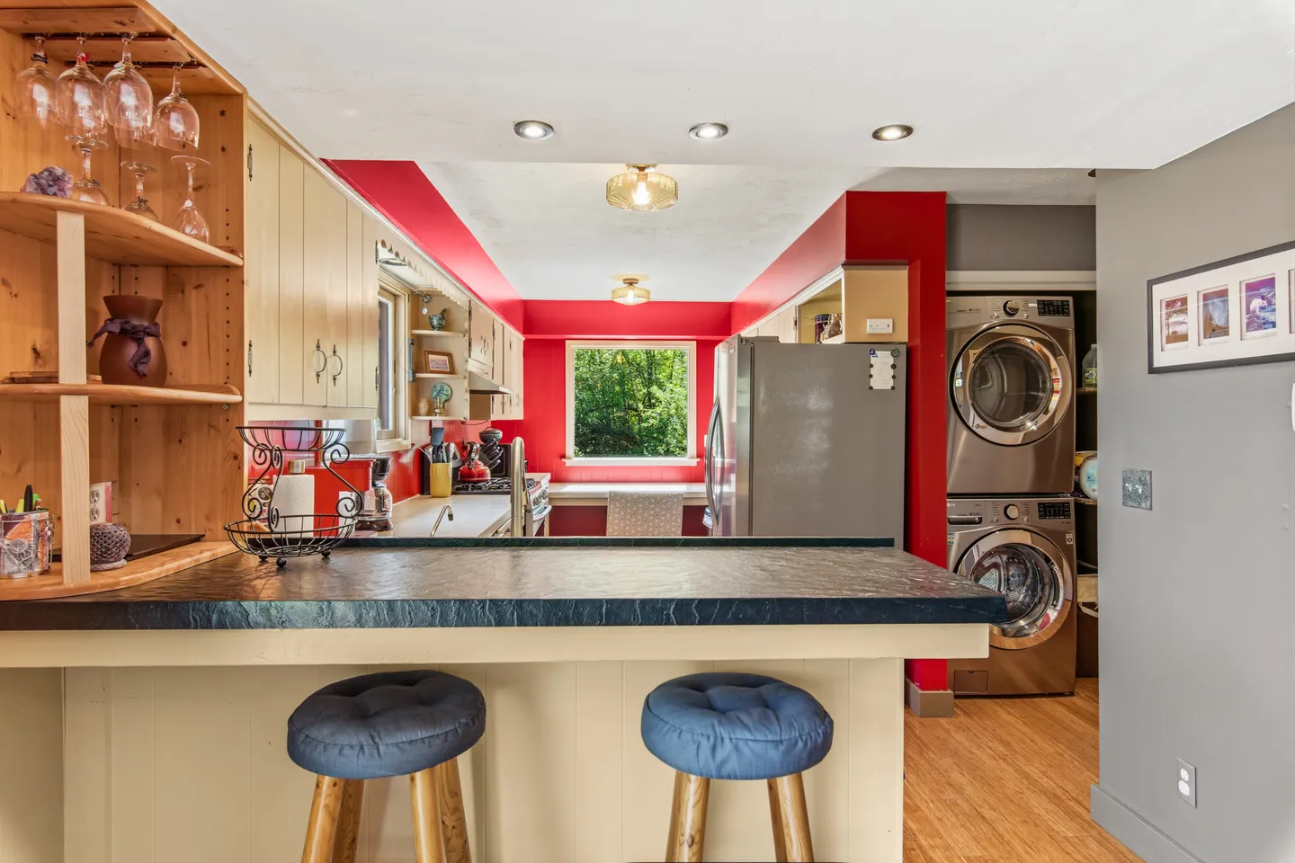 A kitchen with a dark-topped breakfast bar with two blue barstools, light wood cabinets, red accent walls, and a stacked washer and dryer in view.