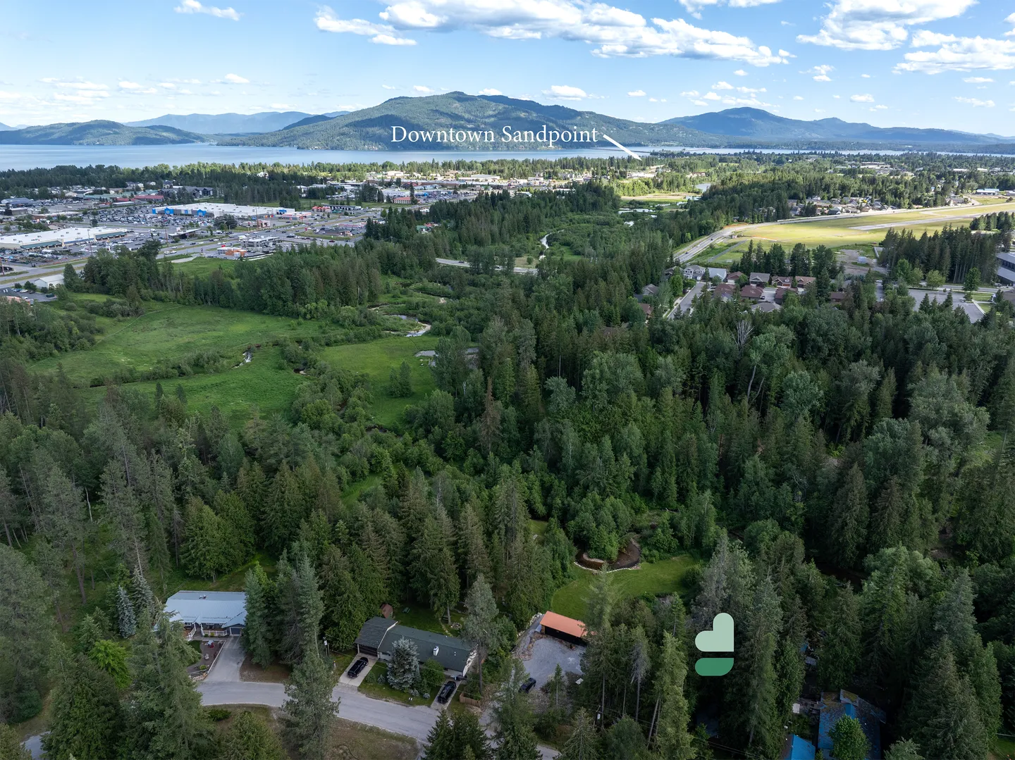 Aerial view of a forested neighborhood with houses nestled in evergreen trees, looking toward a town, lake, and mountains under a blue sky.