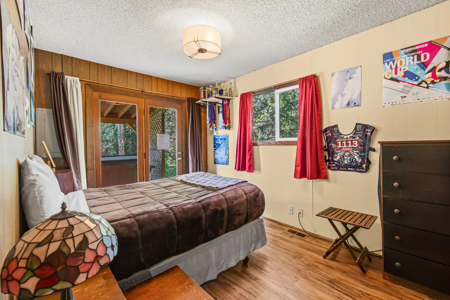 Bedroom with a bed, wood-paneled feature wall, sliding glass doors leading to a covered outdoor area, and a window with red curtains.