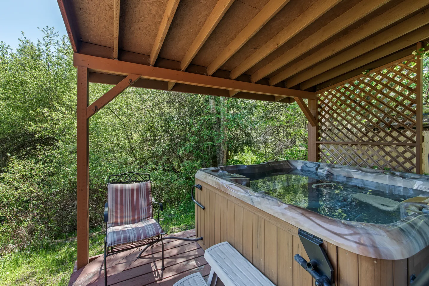 A covered outdoor wooden deck with a filled hot tub, a striped patio chair, and surrounding green trees.