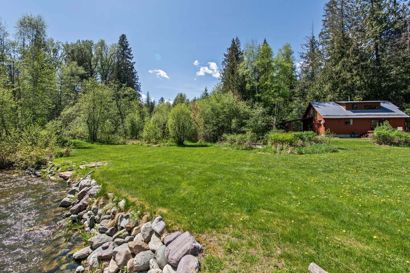 A wide view of a grassy yard bordering a rocky river bank with a house surrounded by tall pine trees in the background.