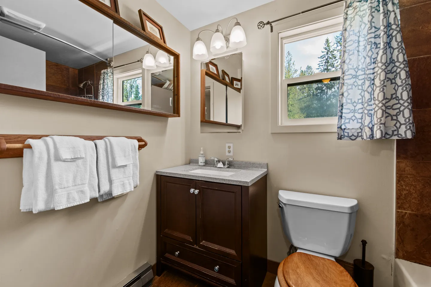 Bathroom featuring a dark wood vanity with a stone countertop, a white toilet with a wooden lid, and a window with a patterned curtain.