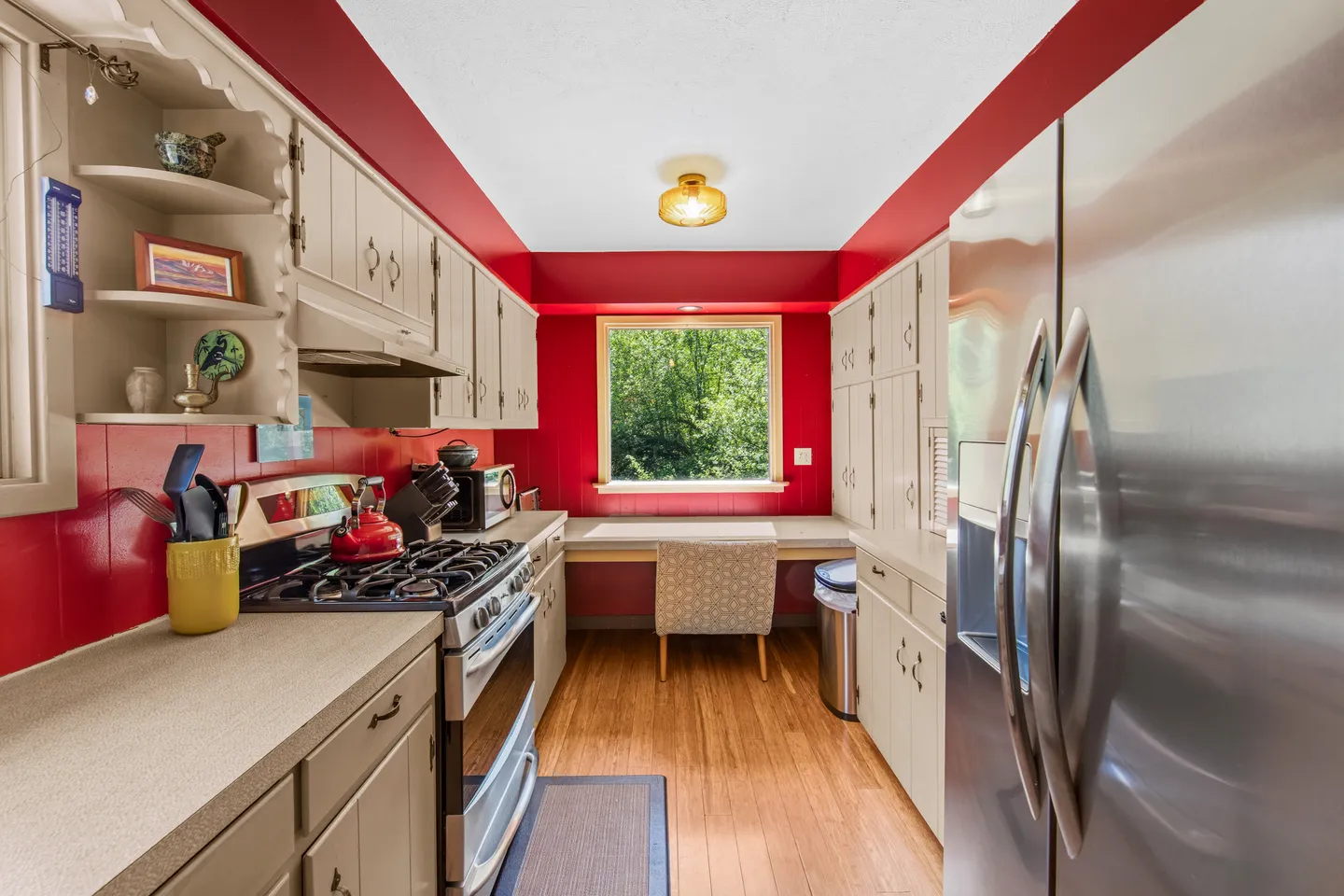 Galley kitchen with light cabinetry, bold red walls, stainless steel appliances, and a window desk at the far end.