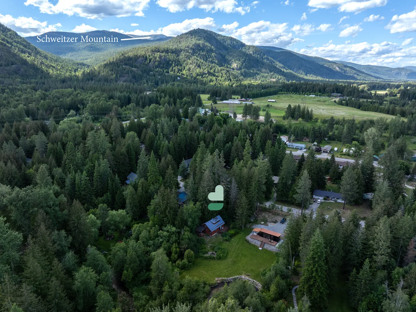 Aerial view of a secluded home surrounded by dense pine trees with large green mountains and blue sky in the background.