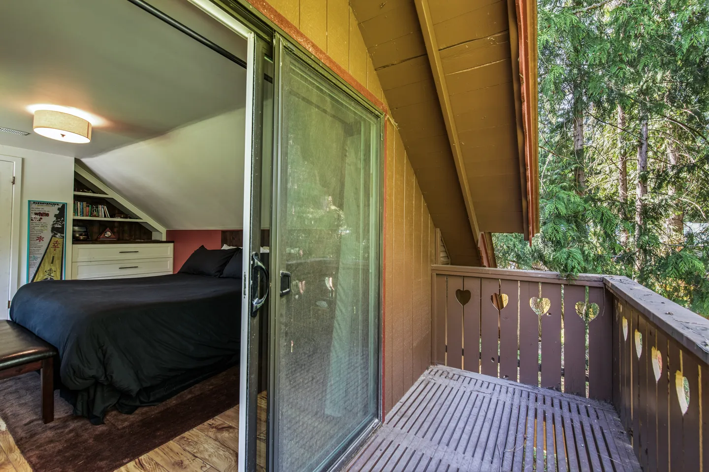 A wooden balcony with heart-shaped cutouts on the railing looks into an adjacent bedroom through a sliding glass door.