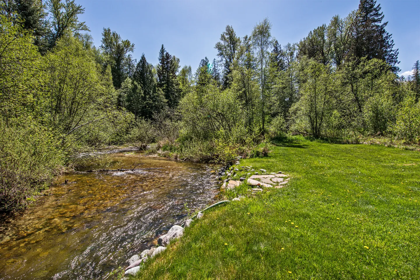 A clear, shallow creek running alongside a grassy lawn lined with stones, surrounded by dense green forest under a blue sky.