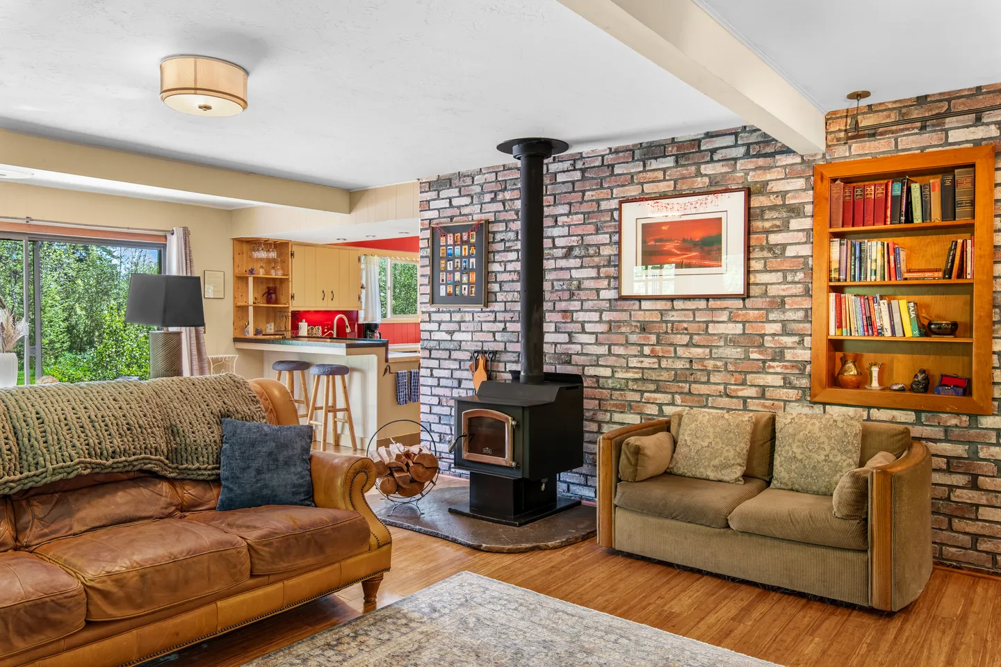 Living room with an exposed brick wall, a black freestanding wood stove, built-in bookshelf, and seating overlooking an open kitchen.