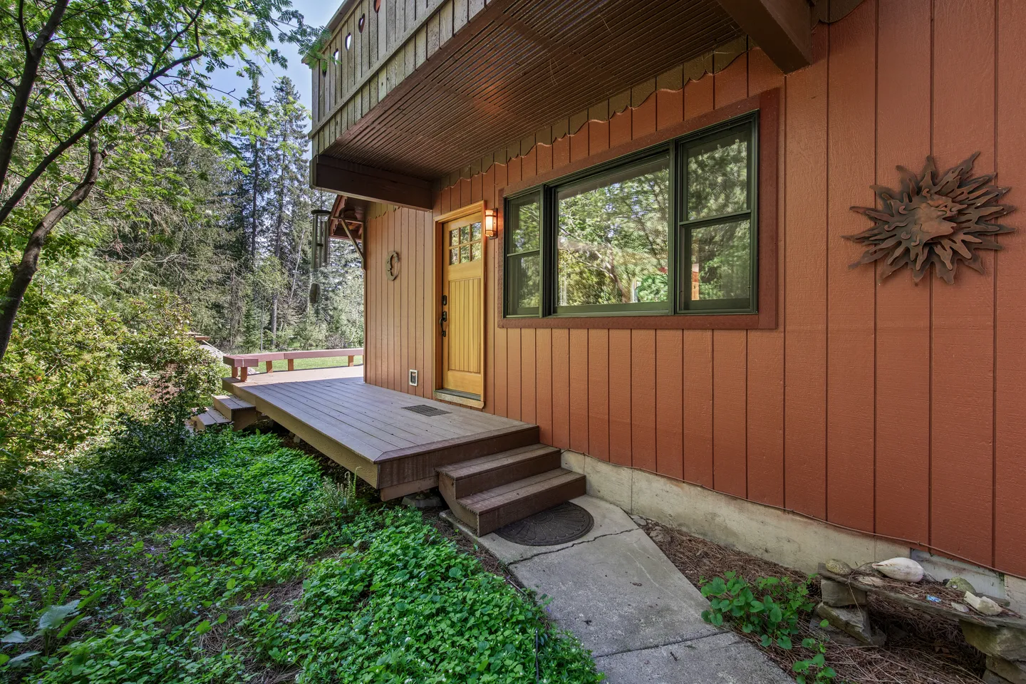 Exterior entrance of a home featuring a wooden walkway, small steps, an orange-paneled facade, a wooden front door, and surrounded by green foliage.